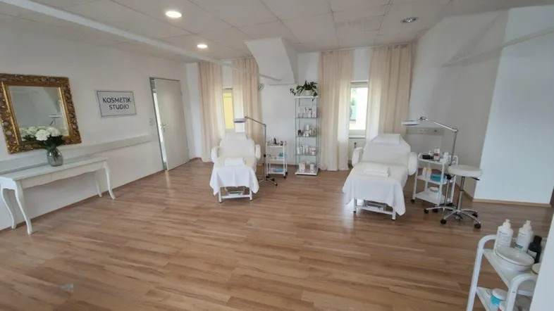 Helles Kosmetikstudio mit zwei Behandlungsstühlen, Holzboden und großen Fenstern für viel Tageslicht.