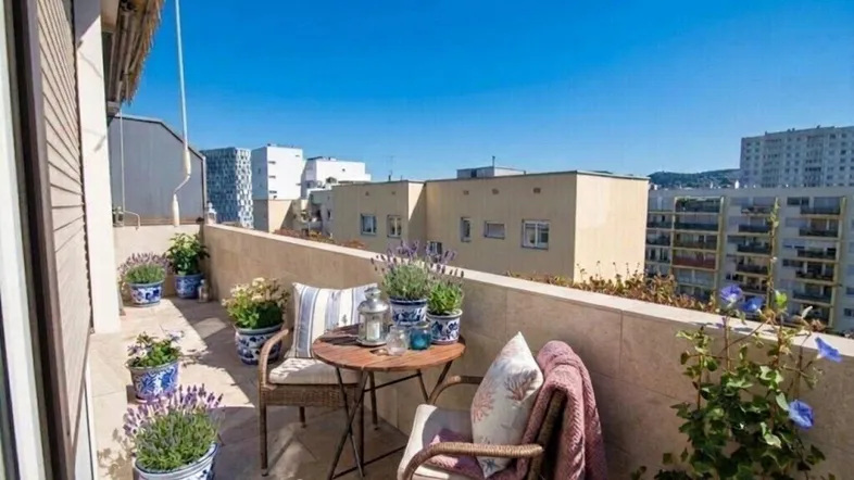 Heller Balkon mit bequemen Sitzgelegenheiten, vielen Pflanzen und Blick auf die umliegenden Gebäude.