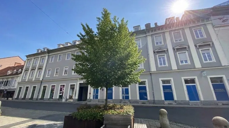 Historisches Gebäude mit grauer Fassade und Geschäften im Erdgeschoss unter blauem Himmel.