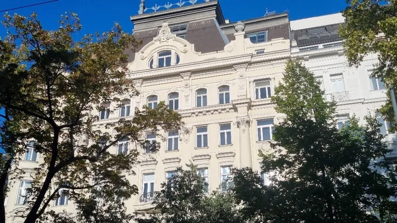 Klassische Gebäudefassade mit vielen Fenstern und Bäumen im Vordergrund unter blauem Himmel.