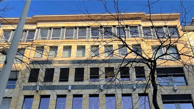 Gebäudefassade mit vielen Fenstern und klarem Himmel, umgeben von kahlen Bäumen, die das Licht filtern.