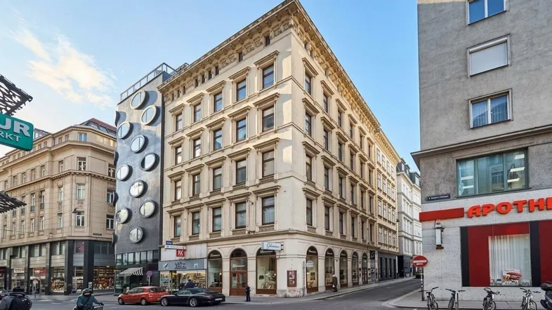 Historisches Gebäude an einer belebten Straßenecke mit klassischer Architektur und angrenzendem modernen Bau.