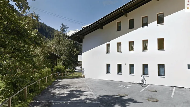 Weißes Mehrfamilienhaus mit Satteldach, Parkplätzen und Bergblick in grüner Umgebung.