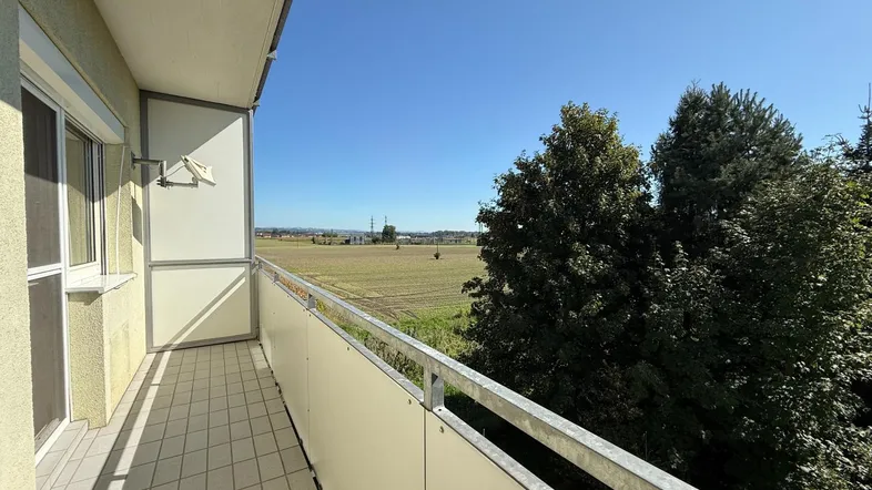 Heller Balkon mit Fliesenboden und weitem Blick über grüne Felder und Bäume.