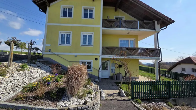 Gelbes Einfamilienhaus mit Satteldach, Balkonen und gepflegtem Vorgarten in ländlicher Umgebung.