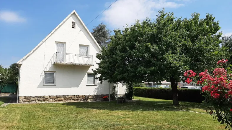 Weißes Einfamilienhaus mit Satteldach, kleinem Balkon und großem Gartenbereich.