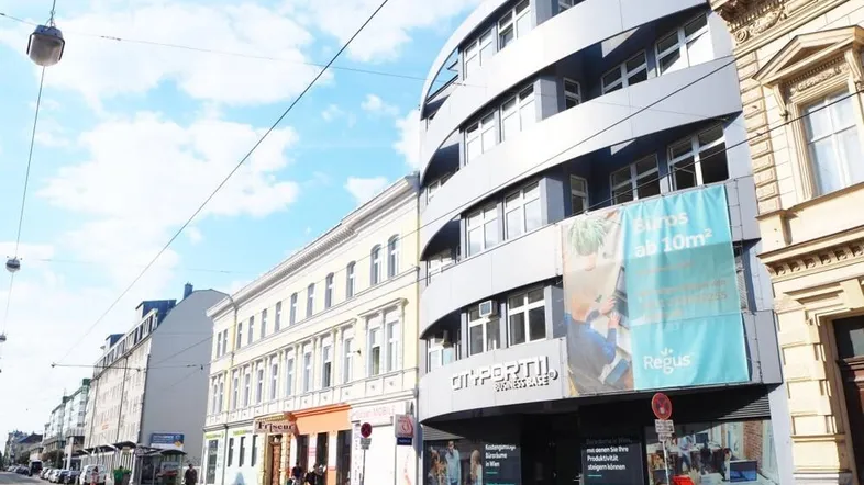 Modernes Geschäftsgebäude mit geschwungener Fassade und großen Fenstern, umgeben von traditioneller Architektur an einer belebten Straße.