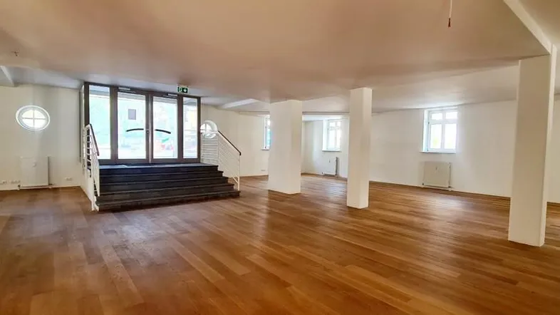 Großer, offener Raum mit hellem Holzfußboden, weißen Wänden und einer breiten Treppe zum Eingangsbereich.
