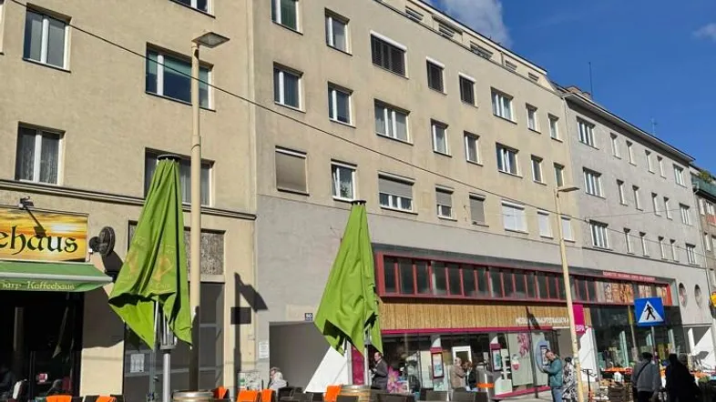 Gebäudeansicht mit Straßencafé und grünen Sonnenschirmen an einem sonnigen Tag.
