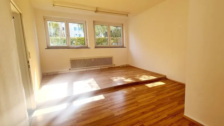 Heller, leerer Raum mit Holzfußboden und zwei Fenstern, die viel Tageslicht hereinlassen.