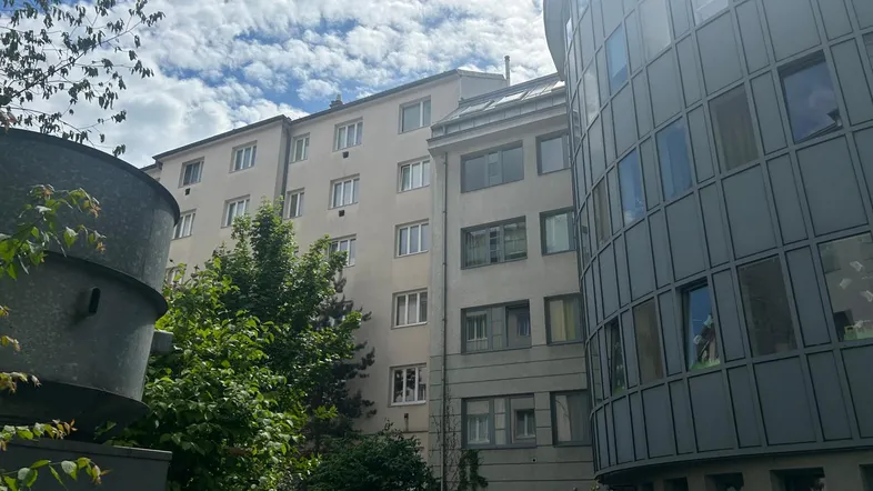 Ansicht von zwei Gebäuden, eines davon mit einer modernen, geschwungenen Fassade, umgeben von Bäumen und Grünflächen.
