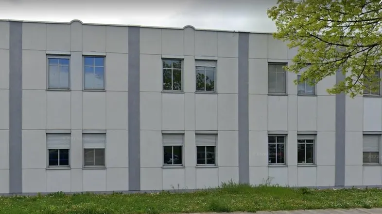 Graue Fassade eines Bürogebäudes mit mehreren Fenstern und Jalousien, umgeben von Gras und Bäumen.