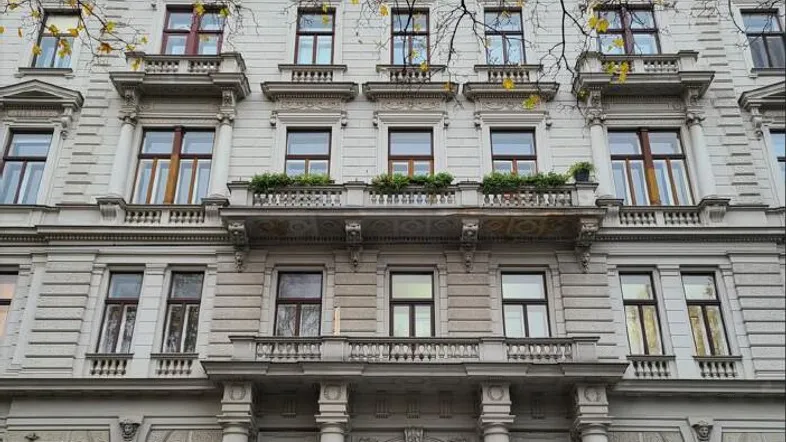 Prächtige Fassade eines Altbaus mit vielen Fenstern und verzierten Balkonen, umgeben von Herbstlaub.