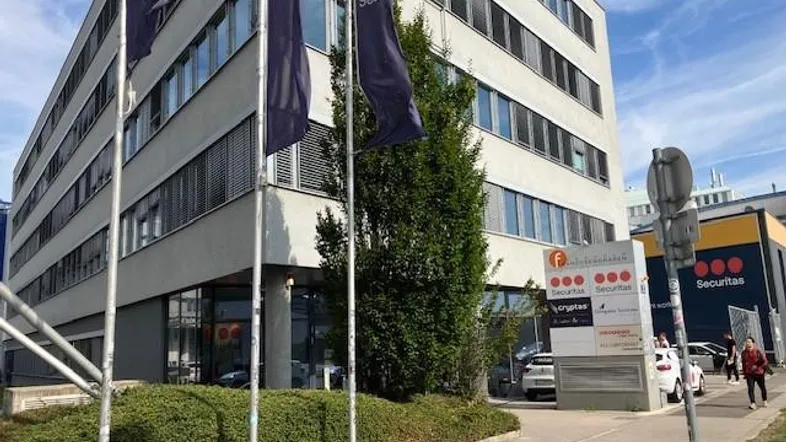 Modernes Bürogebäude mit mehreren Etagen und großen Fenstern, umgeben von Grünflächen und einer Straße.