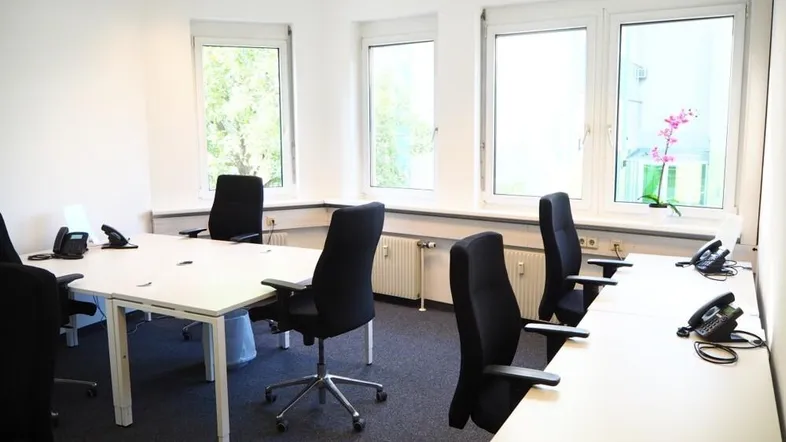Helles und modernes Büro mit mehreren Arbeitsplätzen, großen Fenstern und Teppichboden für eine angenehme Arbeitsatmosphäre.