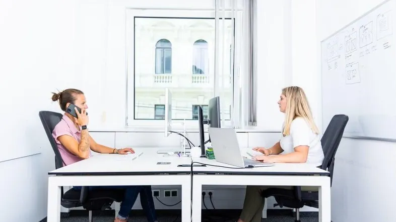 Zwei Personen arbeiten an Schreibtischen in einem hellen Büro mit Blick auf ein historisches Gebäude.