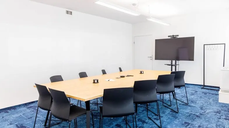 Moderner Konferenzraum mit großem Holztisch, schwarzen Stühlen und einem großen Bildschirm für Präsentationen.