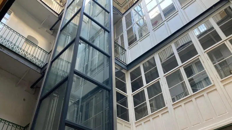 Innenhof eines Gebäudes mit einem modernen Glasaufzug und umlaufenden Balkonen auf mehreren Etagen.