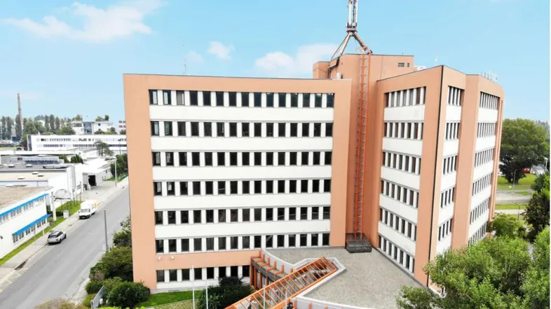 Modernes Bürogebäude mit mehreren Etagen und einer Antenne auf dem Dach, umgeben von Grünflächen und einer Straße.