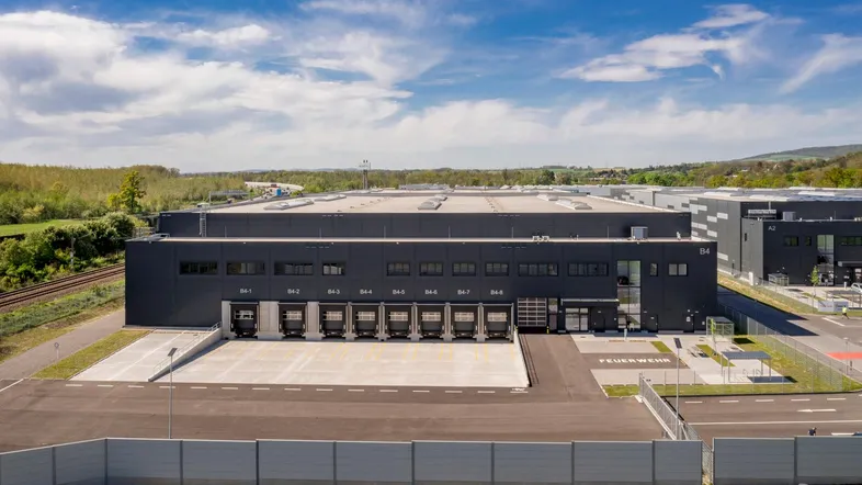 Luftaufnahme eines modernen Logistikzentrums mit zahlreichen Laderampen und weitläufigen Zufahrtswegen unter blauem Himmel.