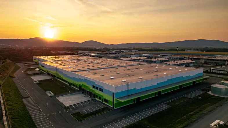 Große moderne Lagerhalle mit farbiger Fassade bei Sonnenuntergang, umgeben von Bergen und Landschaft.
