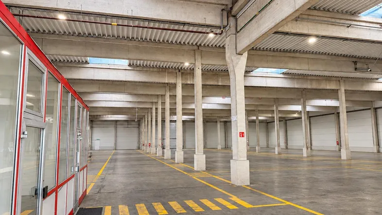 Geräumige Lagerhalle mit hohen Decken, Betonstützen und gelben Bodenmarkierungen, sowie einem abgetrennten Bürobereich.