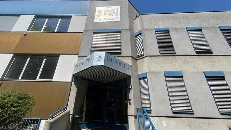 Eingangsbereich eines Gebäudes mit blauen Akzenten, grauer Fassade und großen Fenstern unter blauem Himmel.