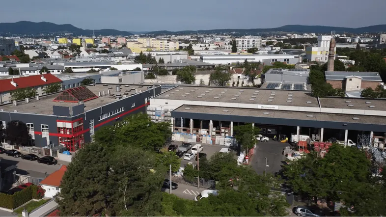 Luftaufnahme eines Gewerbegebiets mit mehreren Gebäuden und umliegender Stadtlandschaft unter blauem Himmel.