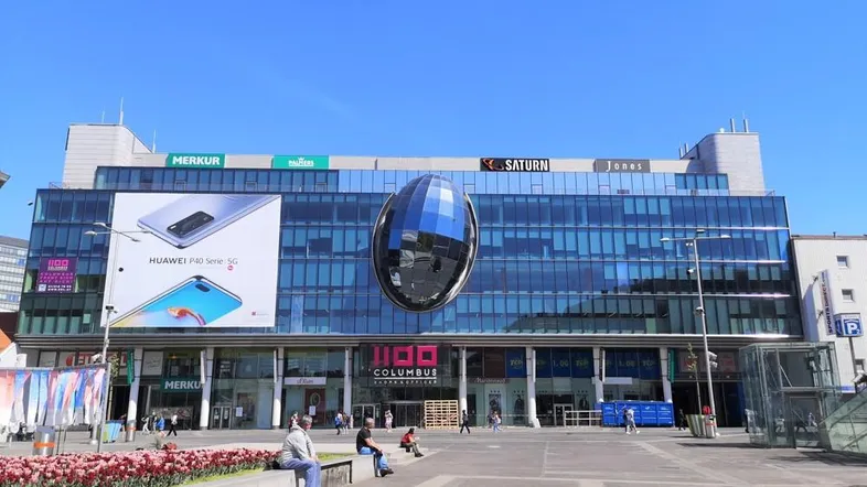 Großes modernes Einkaufszentrum mit Glasfassade und Werbetafeln unter blauem Himmel.