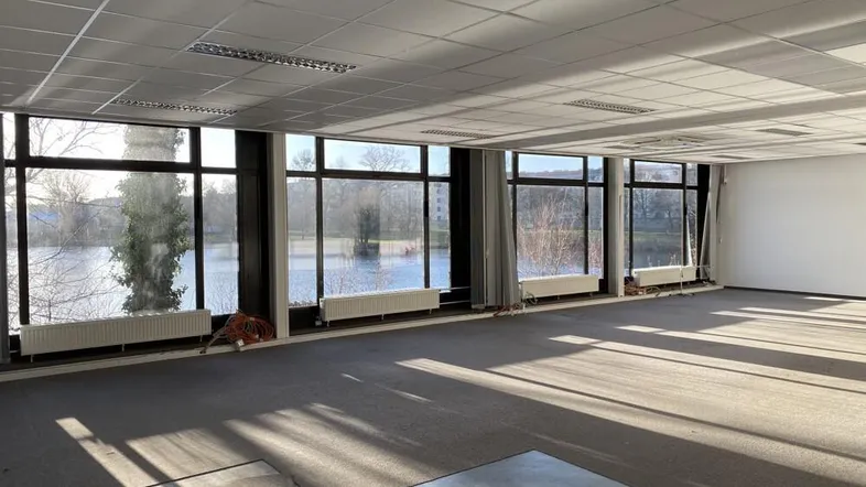 Geräumiger Büroraum mit großen Fenstern und Blick auf einen See, ausgestattet mit Teppichboden und Heizkörpern.