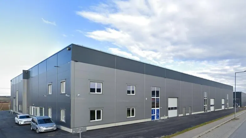 Modernes Industriegebäude mit grauer Fassade, großen Fenstern und Glasfronten unter bewölktem Himmel.