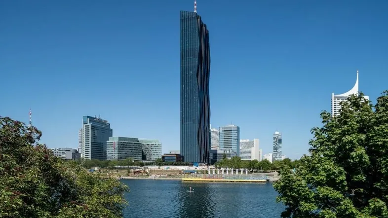Moderner Wolkenkratzer am Flussufer mit blauer Glasfassade und umliegenden Gebäuden unter klarem Himmel.