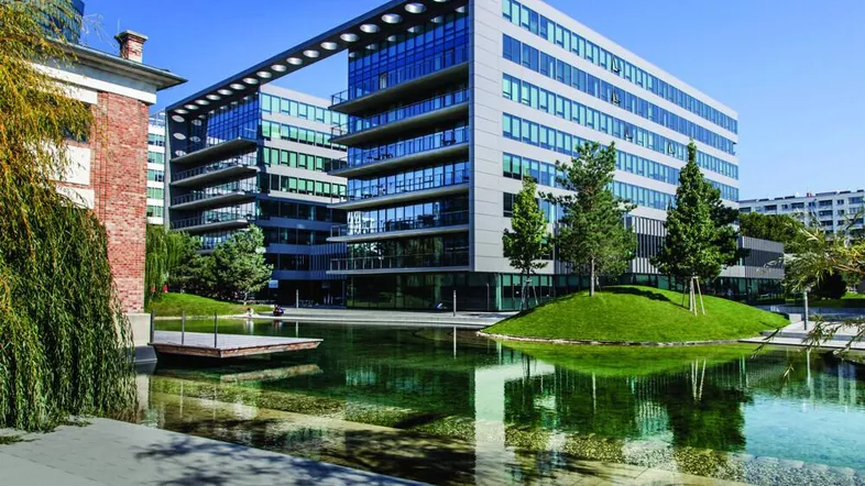Modernes Bürogebäude mit Glasfassade, umgeben von einem Wasserbecken und grünen Inseln.