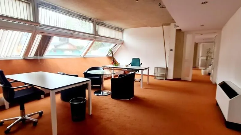 Helles Büro mit großen Fenstern, Teppichboden und mehreren Arbeitsplätzen unter einem Dachschrägenbereich.