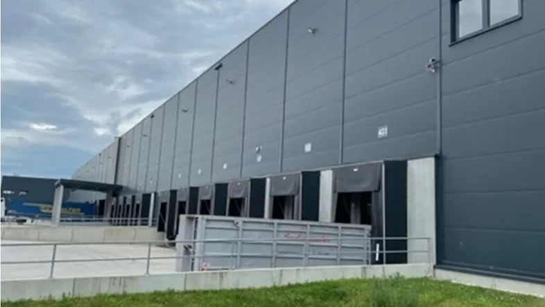 Großes, modernes Industriegebäude mit zahlreichen Laderampen unter bewölktem Himmel und Grünfläche im Vordergrund.