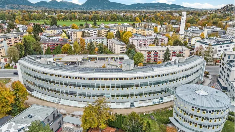 Luftaufnahme eines großen, geschwungenen Bürogebäudes in städtischer Umgebung mit Bergen im Hintergrund.