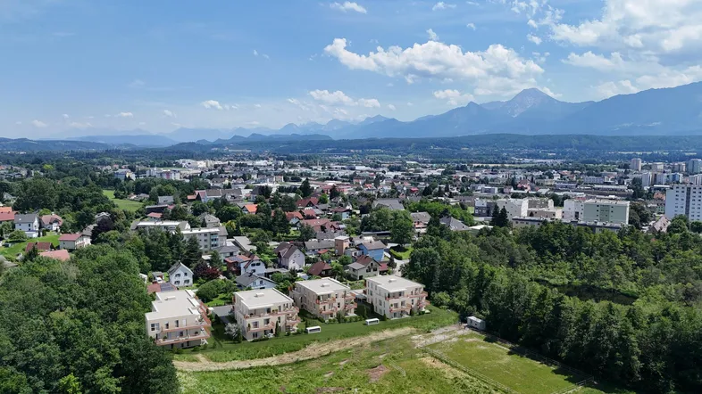 Luftaufnahme einer Wohnsiedlung mit Neubauten und Bergpanorama im Hintergrund unter blauem Himmel.