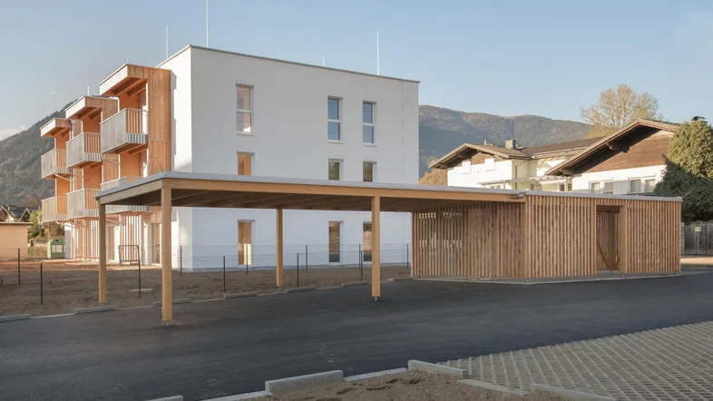 Moderne Wohnanlage mit Holzfassadenelementen, Carport und Parkplätzen vor Bergkulisse.