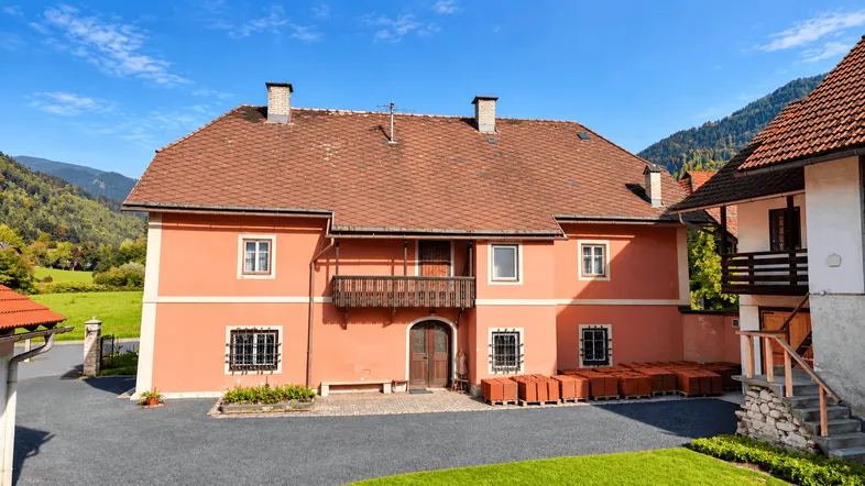 Traditionelles Gebäude mit roter Fassade und Holzbalkon, umgeben von grünen Hügeln unter blauem Himmel.