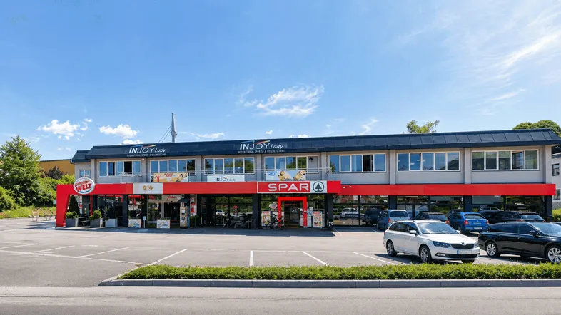 Breite Ansicht eines modernen Geschäftsgebäudes mit Supermarkt und Parkplätzen unter blauem Himmel.