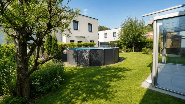 Gepflegter Garten mit Rasenfläche, Bäumen und einem Aufstellpool vor modernen Wohnhäusern.