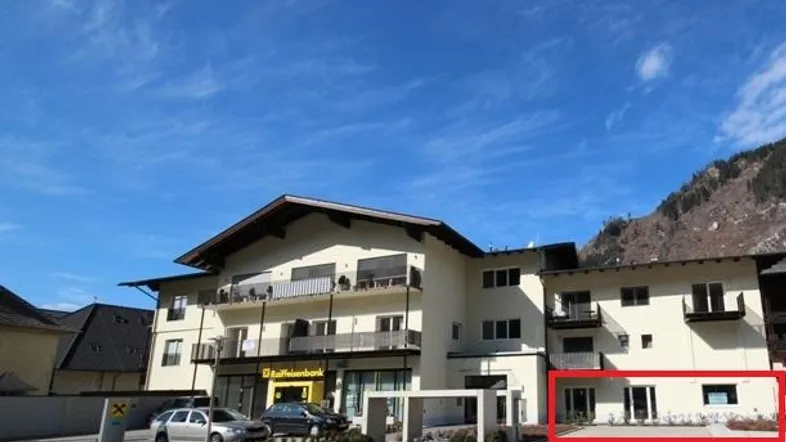 Mehrstöckiges Gebäude mit Balkonen und Geschäftsräumen im Erdgeschoss, umgeben von Bergen unter blauem Himmel.