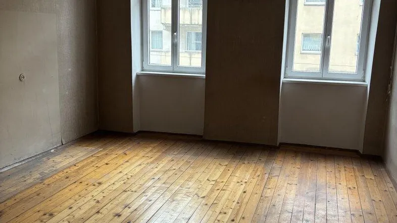 Geräumiges Zimmer mit originalem Holzdielenboden und zwei großen Fenstern, bereit zur Renovierung.