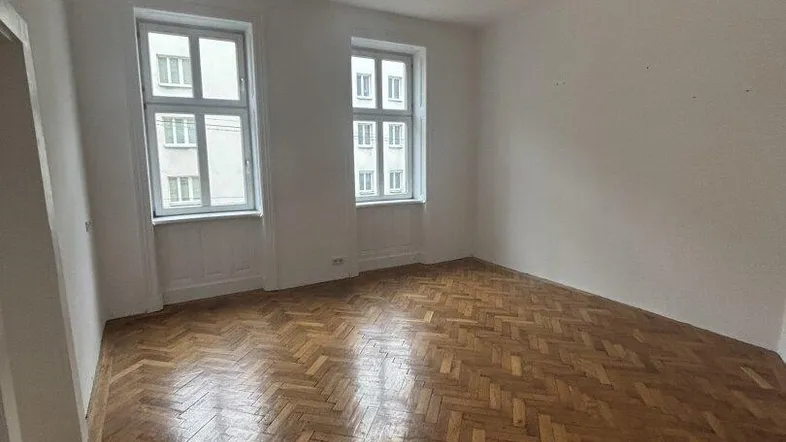 Helles Zimmer mit Fischgrätparkett und zwei großen Fenstern, bereit für die individuelle Gestaltung.