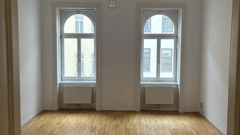 Helles Zimmer mit Parkettboden, weißen Wänden und zwei hohen Bogenfenstern.