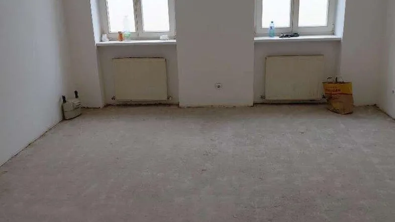 Heller Raum mit zwei Fenstern, Heizkörpern und unfertigem Boden, bereit für die Renovierung.