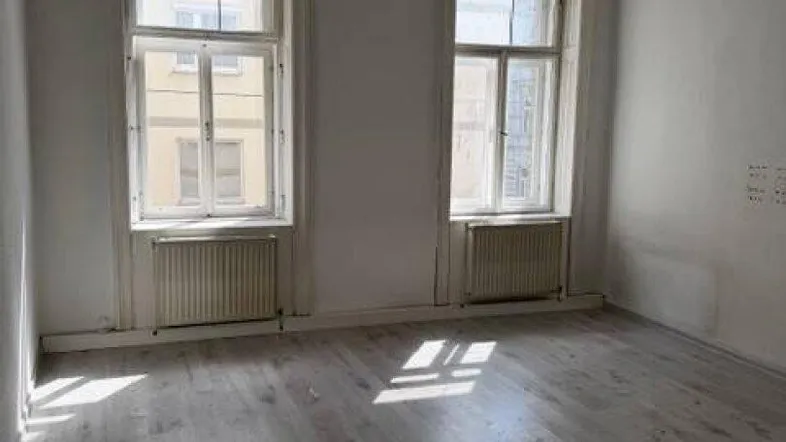 Helles Zimmer mit Laminatboden und zwei großen Fenstern, die viel Tageslicht hereinlassen.