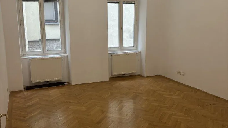 Helles Zimmer mit Fischgrätparkettboden und zwei hohen Fenstern, die viel Tageslicht hereinlassen.