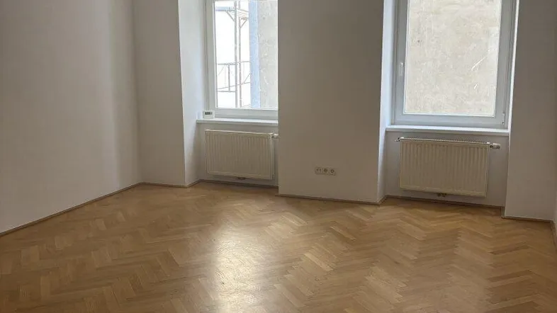 Helles Zimmer mit Fischgrätparkettboden und zwei großen Fenstern, die viel Tageslicht hereinlassen.