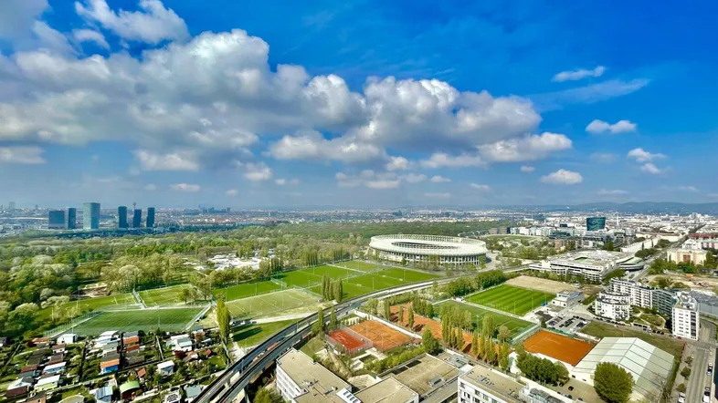 Panoramablick über die Stadt mit Stadion, Donau und umliegenden Wohngebieten unter blauem Himmel.